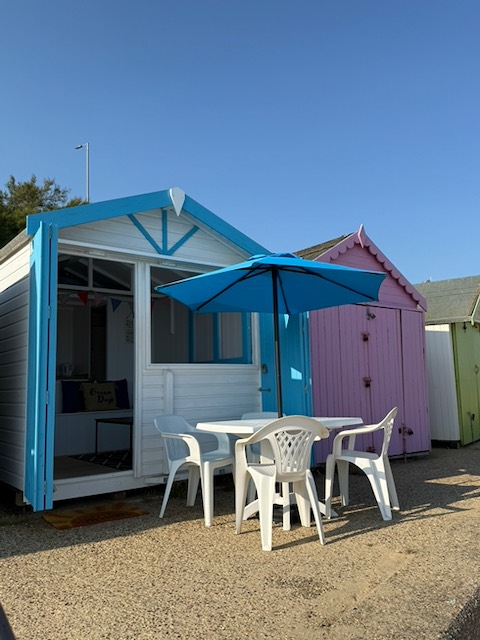 Ocean Days beach hut with its blue bi-fold doors open, a table, chairs and parasol set up outside under a clear blue sky.