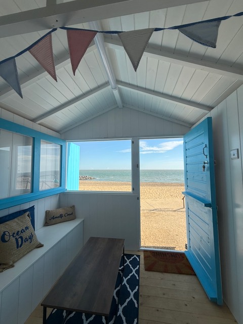 Looking out through the hut's open blue door across the sand to the sea, with bunting overhead and cushions on the bench.
