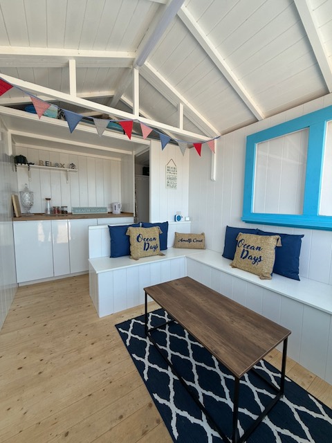 Inside the hut: white bench seating with navy and jute cushions, a table on a navy rug, kitchen units and bunting.